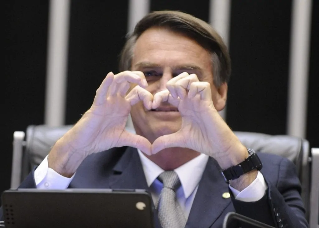 Fazendo sinal de coração com as mãos, o genocida que não adquiriu vacinas a tempo de evitar a morte de pelo menos 95 mil brasileiros.